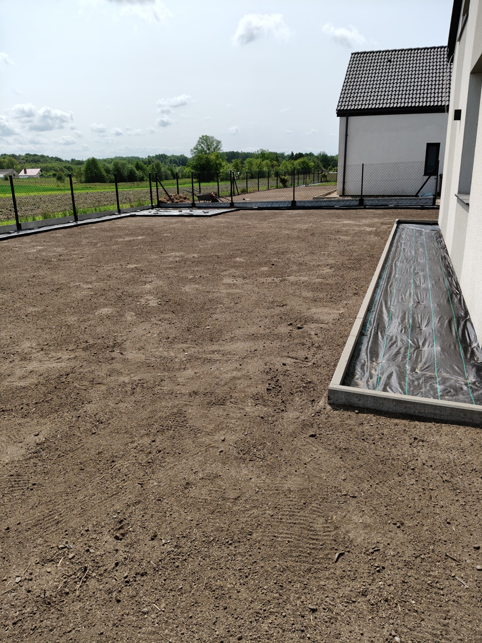 Przygotowany teren pod trawnik z obrzeżami i agrowłókniną przy ścianie domu. Widok na ogrodzenie i zieleń w tle, słoneczny dzień.