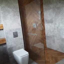 RenovArt - Nowoczesna łazienka z kabiną prysznicową typu walk-in, wykończoną drewnem ułożonym w jodełkę. Szare płytki z dekoracyjnym pasem i podwieszana toaleta.