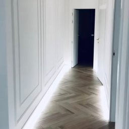 Łukasz Truskolaski - Nowoczesny korytarz z parkietem ułożonym w jodełkę, białe ściany z dekoracyjnymi panelami i subtelnym oświetleniem LED wzdłuż podłogi.
