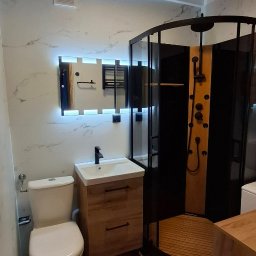 RenovArt - Wykończona łazienka w Głogowie z kabiną prysznicową, toaletą i umywalką z szafką w jasnej kolorystyce. Lustro z podświetleniem LED i marmurowe płytki na ścianach.