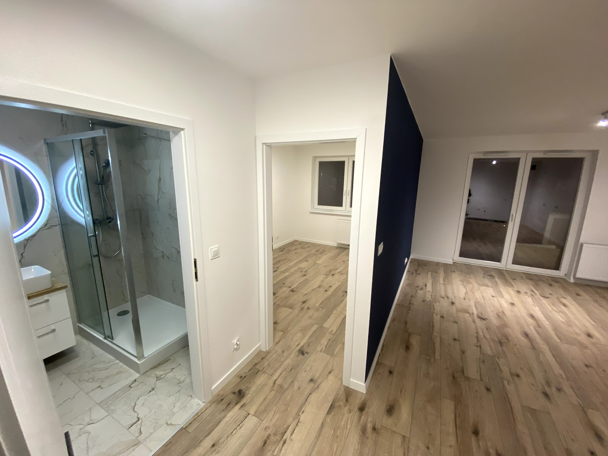 Nowoczesne wnętrze po remoncie: widok na łazienkę z kabiną prysznicową, drewniane panele podłogowe i fragment ściany pomalowanej na granatowo.