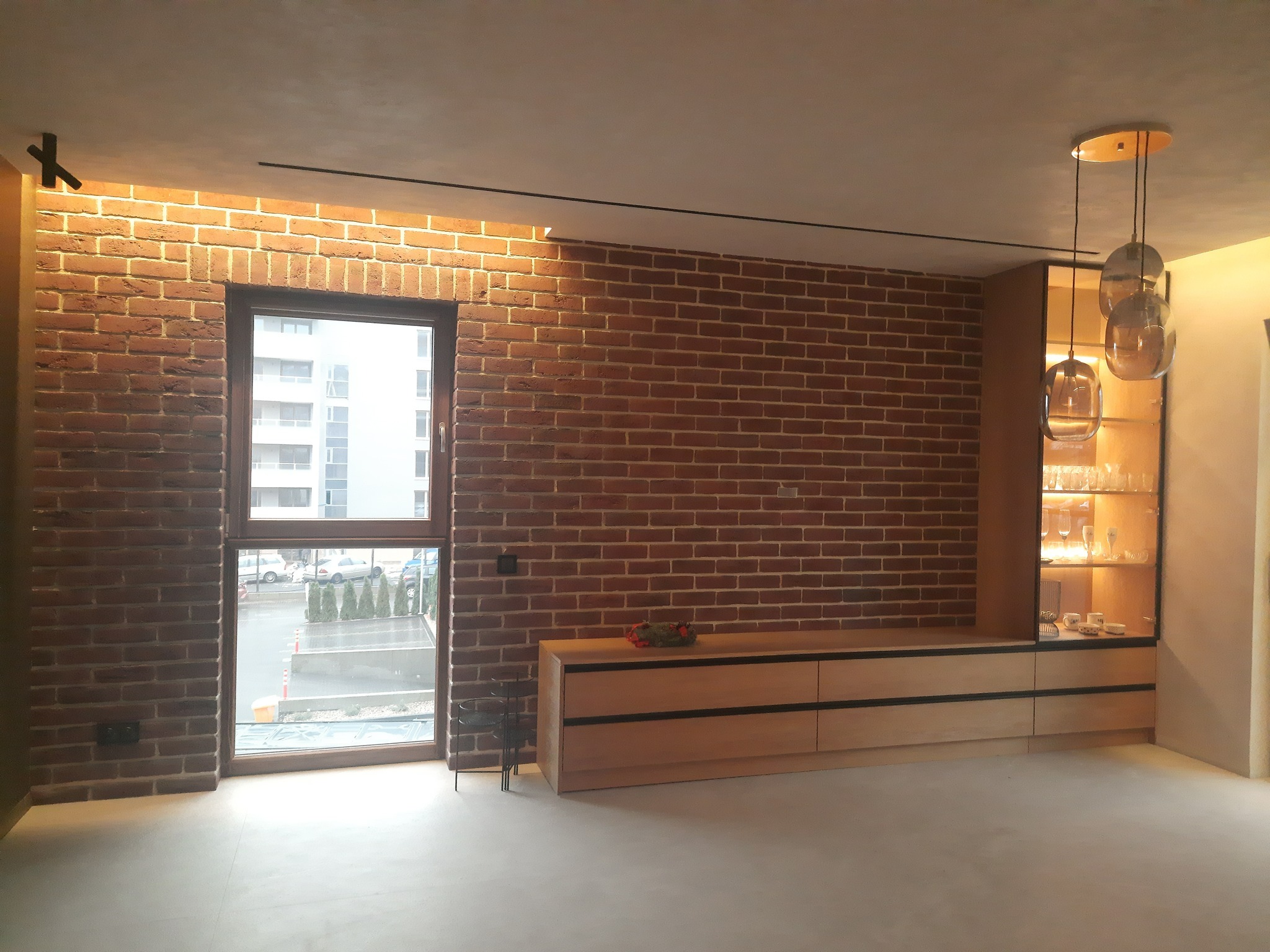 Loftowe wnętrze: ceglana ściana z oknem, drewniana komoda i witryna z oświetleniem punktowym, wiszące lampy. Minimalistyczny design z akcentem industrialnym.