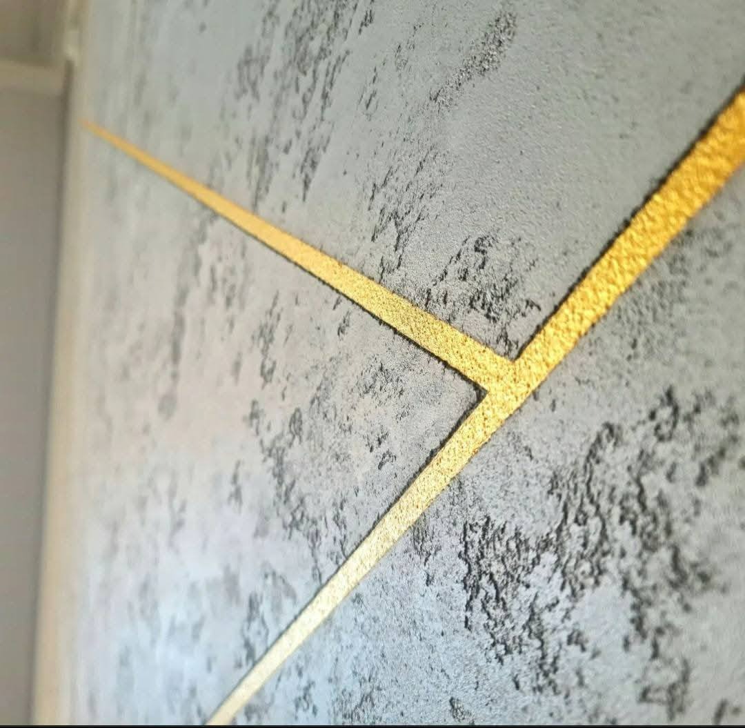 Dekoracyjna ściana z szarą, strukturalną fakturą i złotymi, geometrycznymi liniami tworzącymi wzór. Efektowna aranżacja wnętrza z bliska.