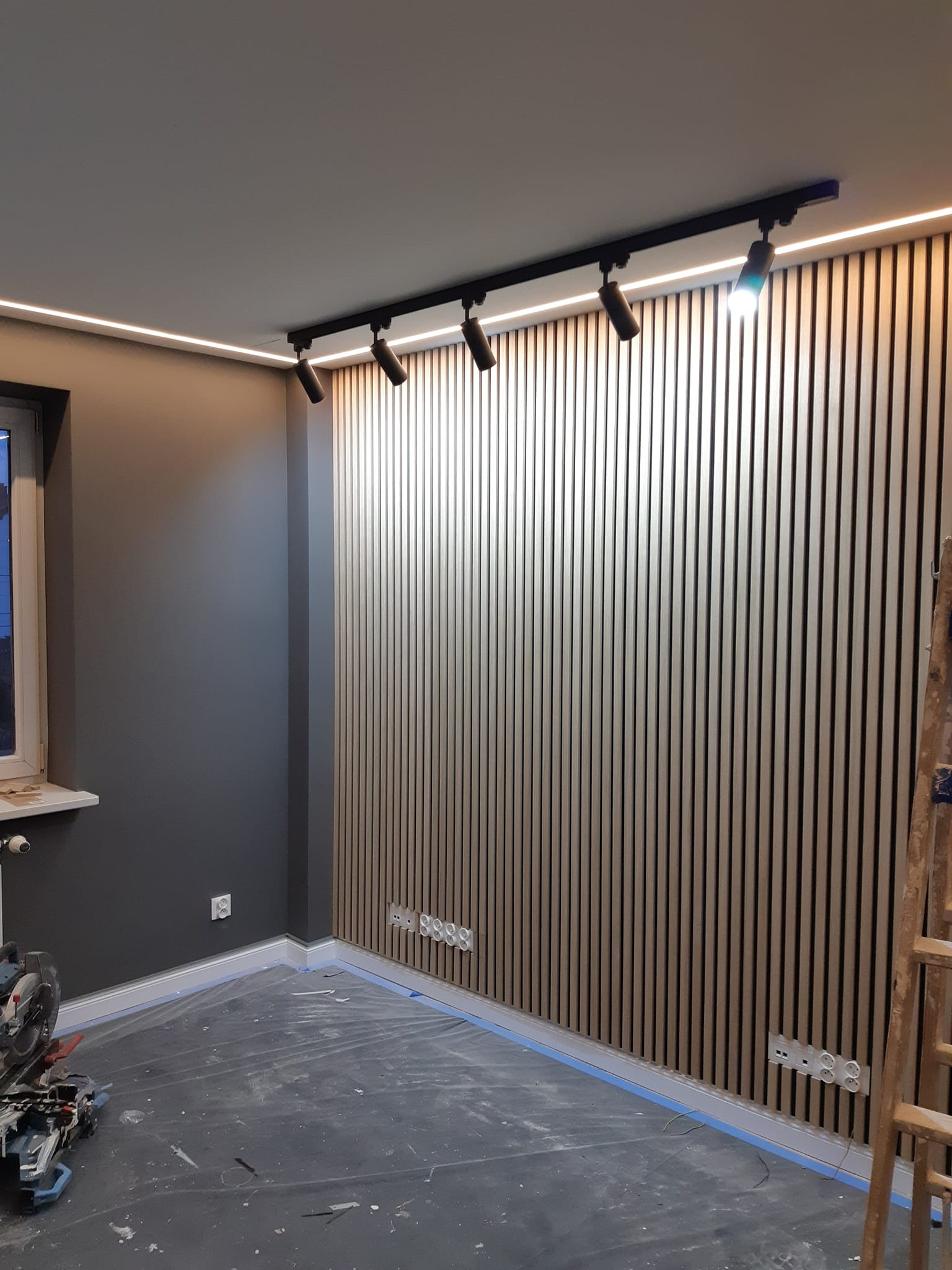 Ściana z drewnianych paneli pionowych podświetlonych ledami i reflektorami, z widocznymi gniazdkami elektrycznymi i elementami wykończenia, w trakcie prac remontowych.