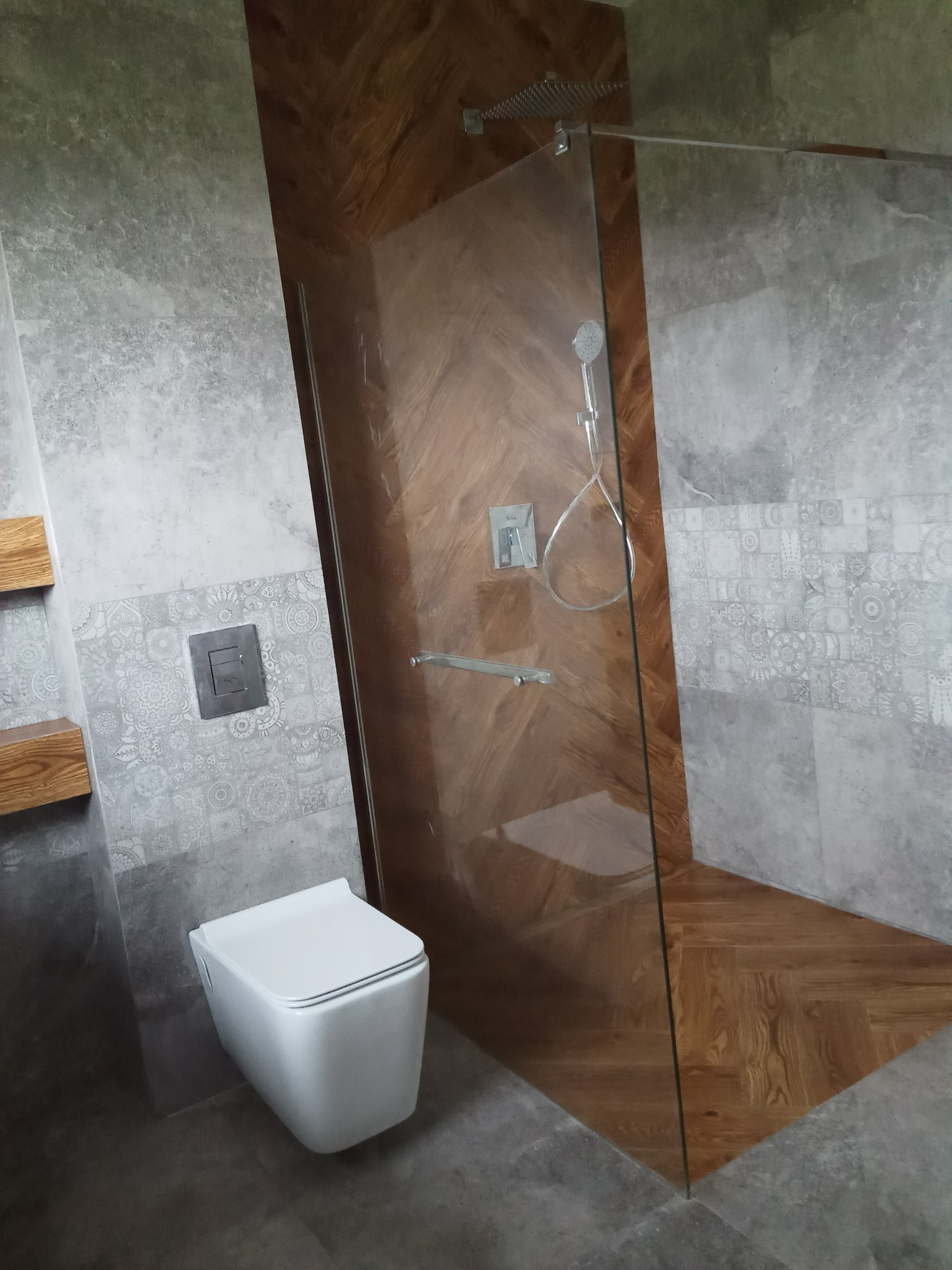 Nowoczesna łazienka z kabiną prysznicową typu walk-in, wykończoną drewnem ułożonym w jodełkę. Szare płytki z dekoracyjnym pasem i podwieszana toaleta.