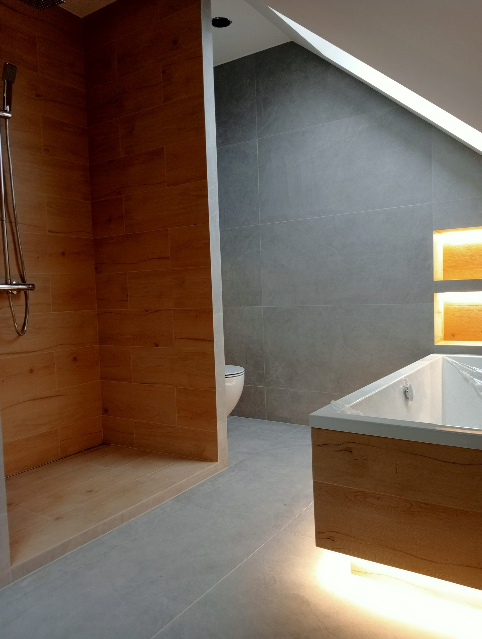 Nowoczesna łazienka na poddaszu: drewnopodobne płytki w strefie prysznica, szare płytki na ścianach, podświetlana wanna. Aranżacja z dbałością o detale.
