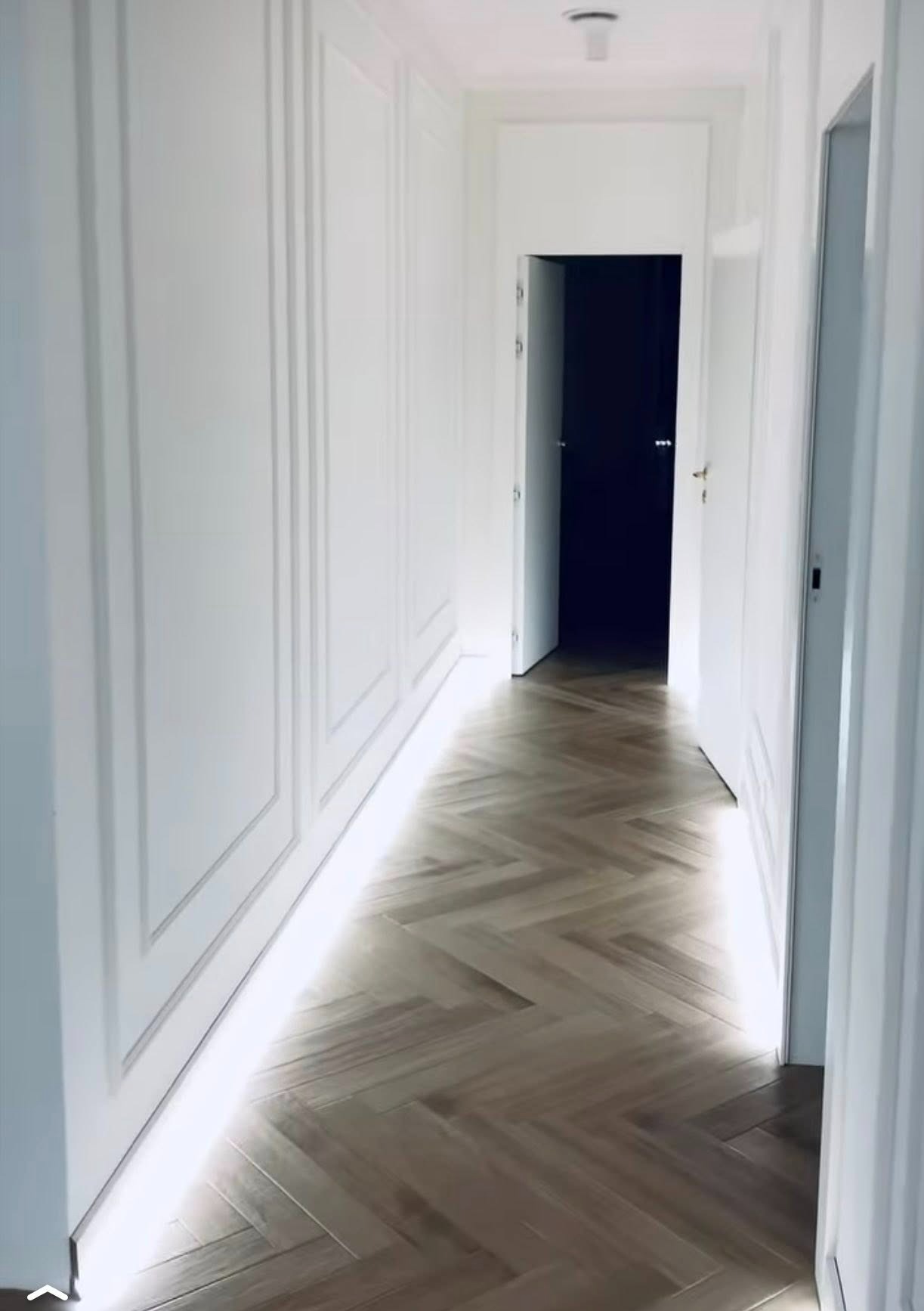 Nowoczesny korytarz z parkietem ułożonym w jodełkę, białe ściany z dekoracyjnymi panelami i subtelnym oświetleniem LED wzdłuż podłogi.