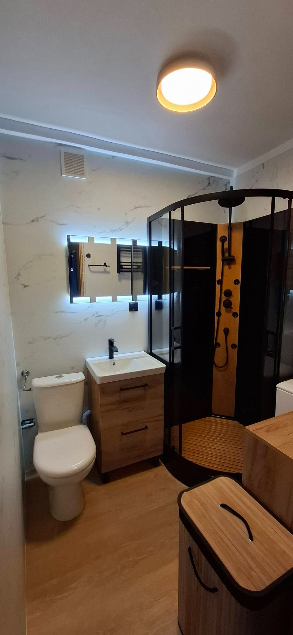 Wykończona łazienka w Głogowie z kabiną prysznicową, toaletą i umywalką z szafką w jasnej kolorystyce. Lustro z podświetleniem LED i marmurowe płytki na ścianach.