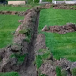 Wykopany rów na trawniku pod system nawadniania. Widoczne fragmenty wykopanej ziemi, trawa oraz drzewa w tle. Etap prac ziemnych przed instalacją systemu.