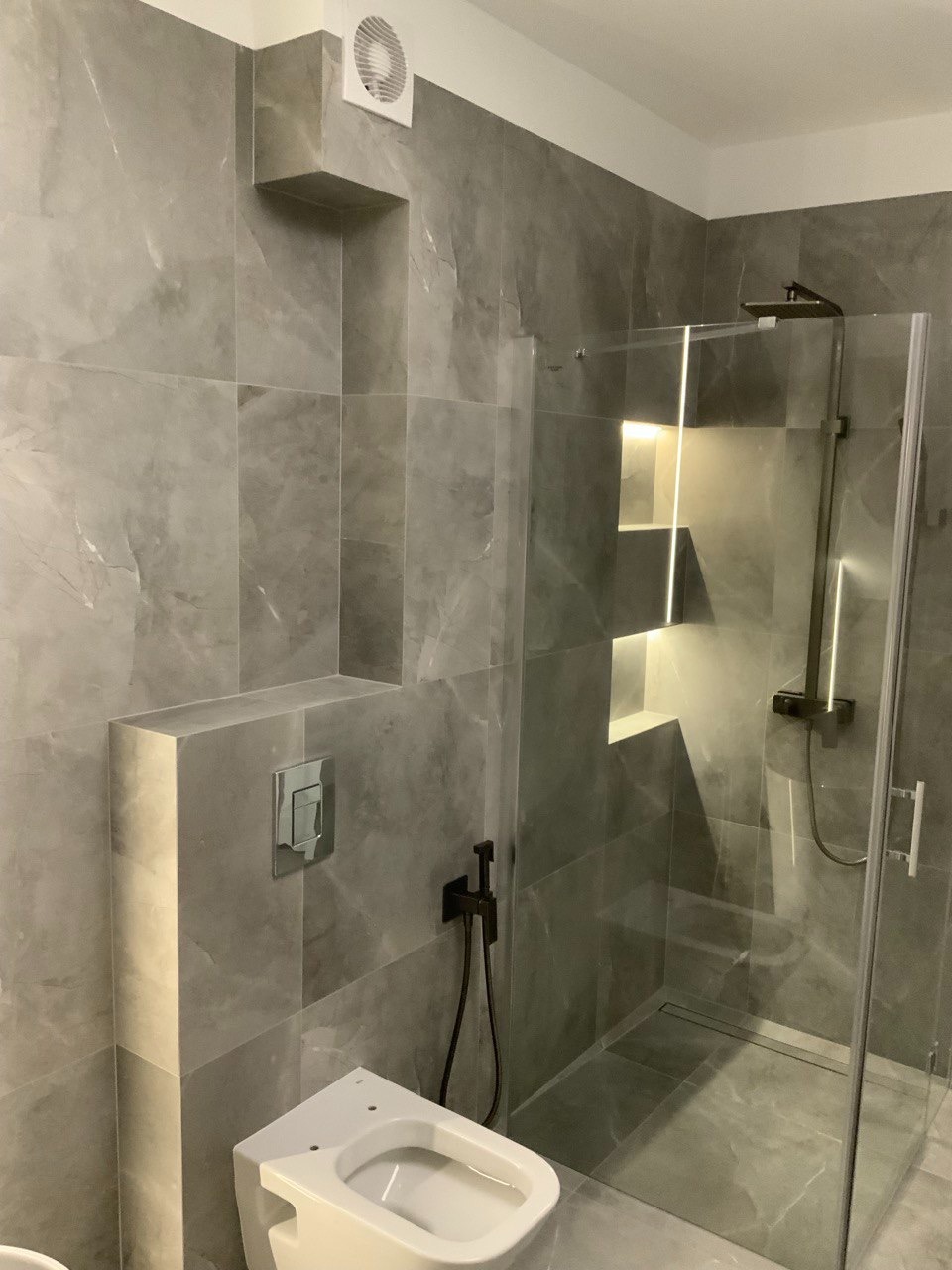 Nowoczesna łazienka z szarymi płytkami imitującymi marmur, kabiną prysznicową typu walk-in i podtynkową toaletą. Widoczna wentylacja i oświetlenie niszowe.