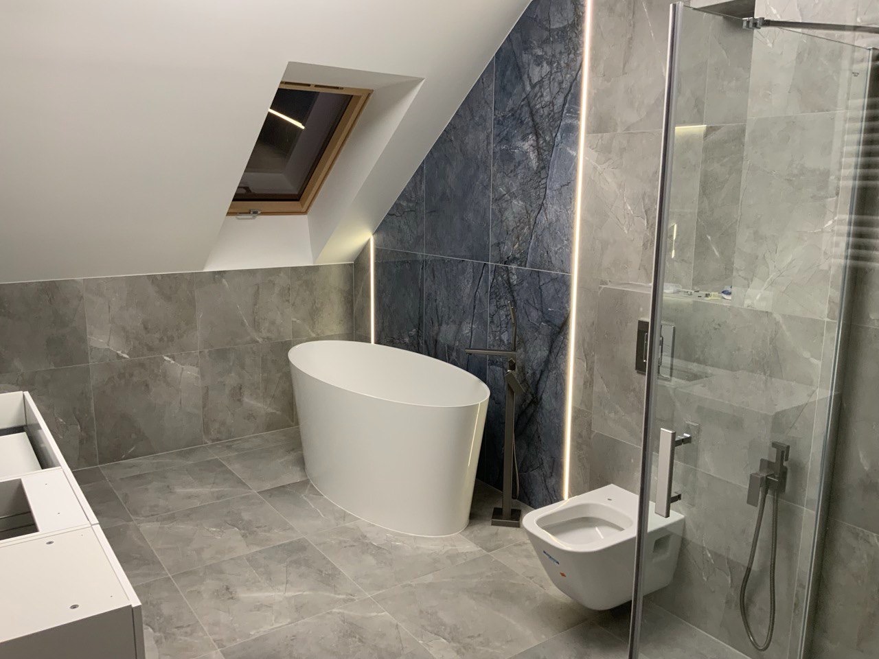 Nowoczesna łazienka na poddaszu z wolnostojącą wanną, szarą marmurową glazurą, oknem dachowym i podświetleniem LED. Kabina prysznicowa ze szklanymi drzwiami.