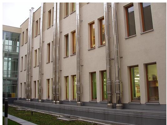 Zewnętrzne, metalowe kominy wentylacyjne zamontowane na elewacji budynku z beżową fasadą i brązowymi ramami okiennymi, widoczne trawnik i kostka brukowa.