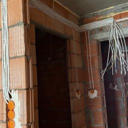ELEKTROKOLBA Instalacje Elektryczne Artur Plebański - Instalacja elektryczna w surowym budynku: kable w peszlach prowadzone po ścianach z cegły, puszki elektryczne w ścianie, przewody zwisające z sufitu.