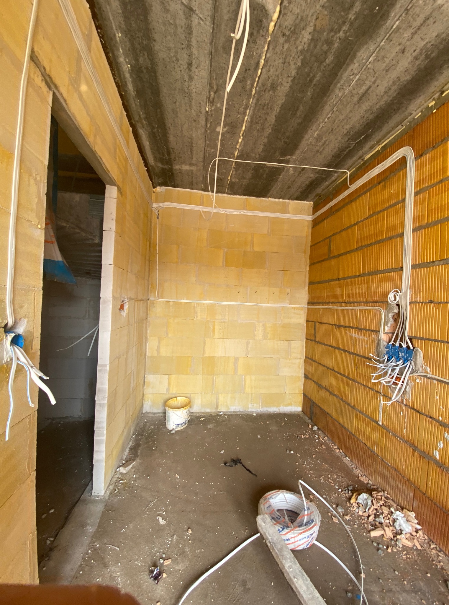 Wnętrze w trakcie prac: widoczne kable elektryczne prowadzone po ścianach z bloczków, puszki instalacyjne i przewody zwisające z sufitu. Na podłodze resztki materiałów budowlanych.