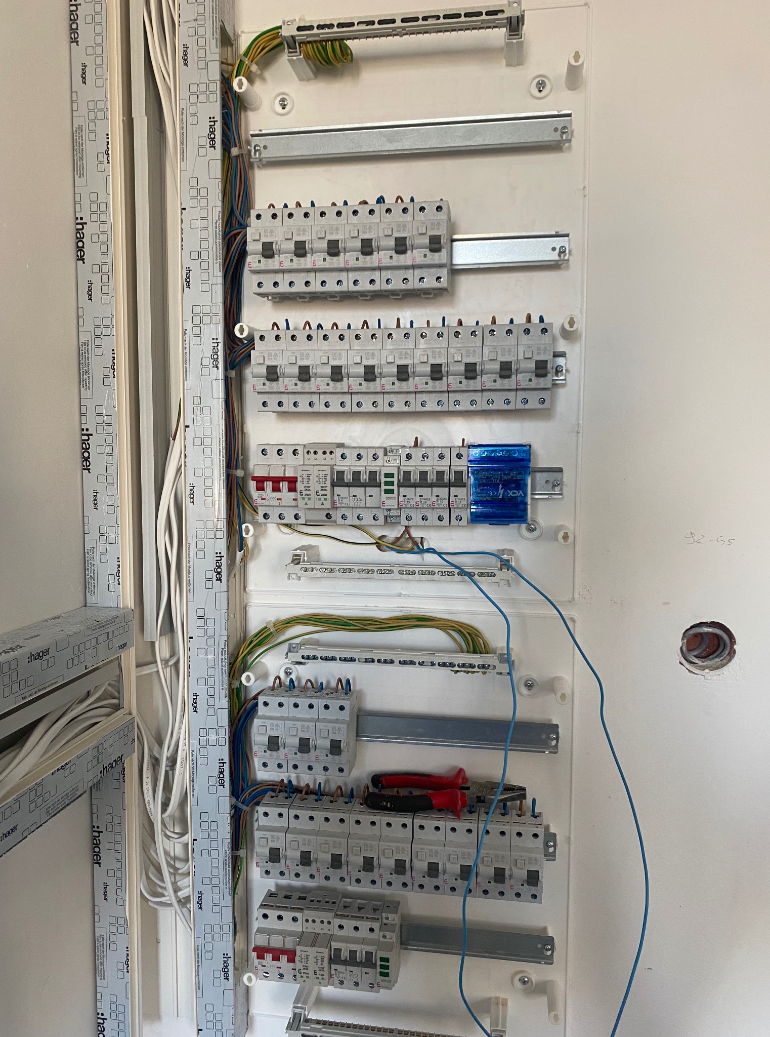Rozdzielnica elektryczna Hager z zainstalowanymi bezpiecznikami, przewodami i narzędziami, widoczna na białej ścianie. Częściowo zdjęta folia ochronna z obudowy.