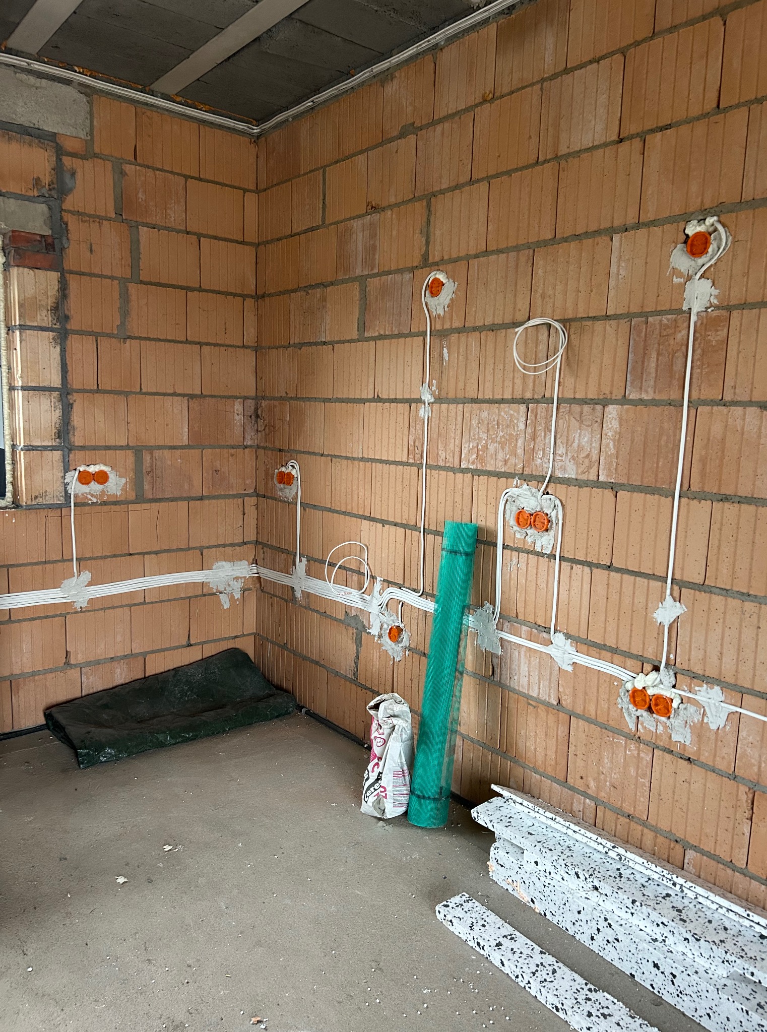 Wnętrze w trakcie prac: widoczne kable elektryczne w ścianach z cegły, puszki instalacyjne z pomarańczowymi elementami, materiały budowlane na podłodze.