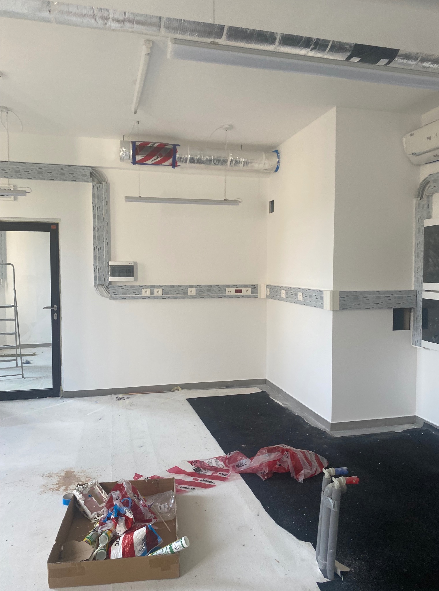 Wnętrze w trakcie prac: szare koryta kablowe na ścianach, gniazdka elektryczne, rury pod sufitem. Na podłodze ślady prac i karton z odpadami.