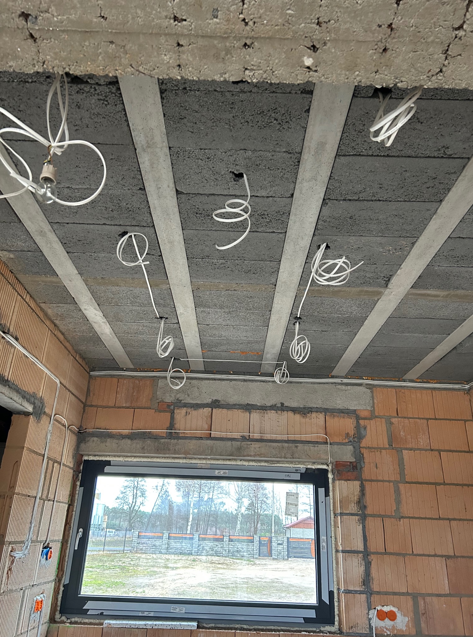 Surowy strop z widocznymi przewodami elektrycznymi, instalacja w trakcie montażu. Ściany z cegły dziurawki, okno z widokiem na zewnątrz.