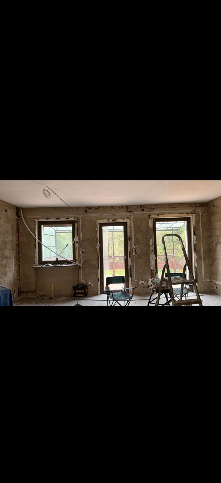 Wnętrze w trakcie remontu z widocznymi przewodami elektrycznymi poprowadzonymi wzdłuż ścian, narzędzia i drabina w pomieszczeniu. Surowy stan ścian.