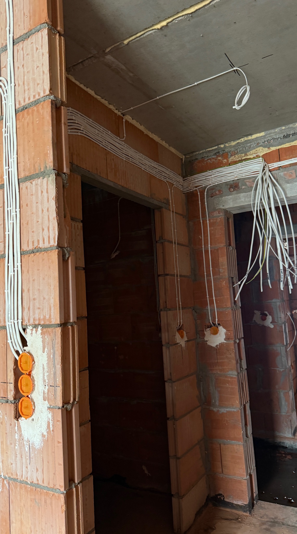 Instalacja elektryczna w surowym budynku: kable w peszlach prowadzone po ścianach z cegły, puszki elektryczne w ścianie, przewody zwisające z sufitu.