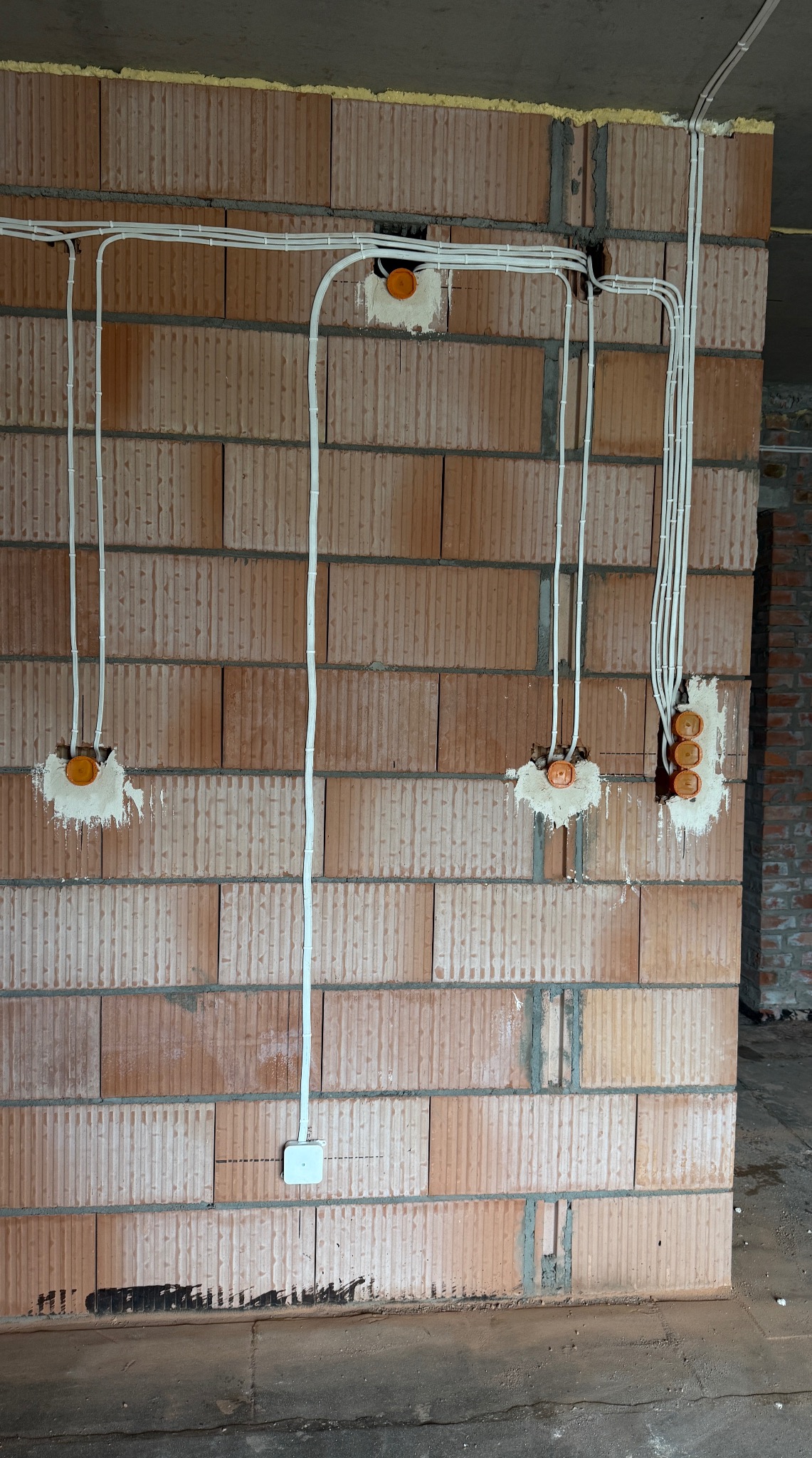 Ściana z pustaków z rozprowadzoną instalacją elektryczną: białe kable prowadzone w bruzdach, puszki pod gniazdka i włączniki, surowy stan wykończenia.