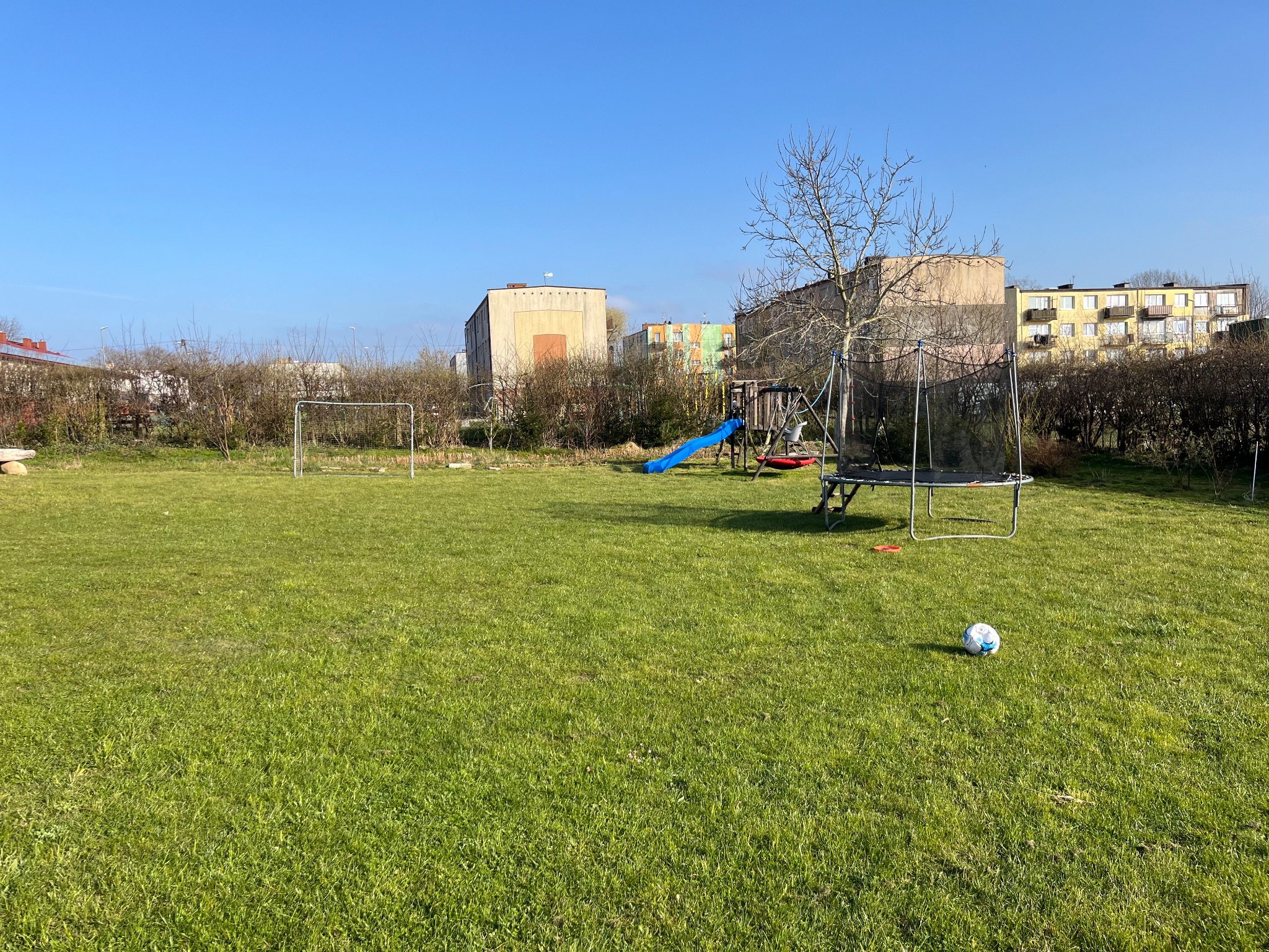 Rozległy trawnik z trampoliną, zjeżdżalnią, bramką piłkarską i piłką. W tle budynki mieszkalne, krzewy i błękitne niebo. Widok na zadbany, choć nieco nierówny trawnik.