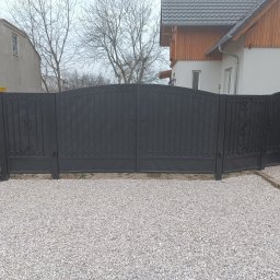 Robstal - Czarna, metalowa brama wjazdowa z dekoracyjnymi elementami, zamontowana przed domem z jasną elewacją i szarym dachem, na podłożu wysypanym białym kamieniem.