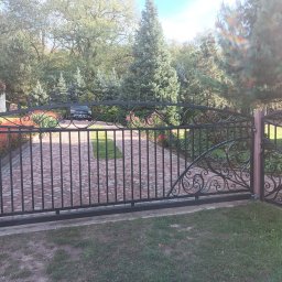 Robstal - Kuta brama wjazdowa z furtką, zdobiona ornamentami. Na podjeździe kostka brukowa, w tle zieleń drzew i krzewów. W oddali zaparkowany czarny samochód.