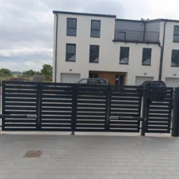 Nowoczesna, przesuwna brama wjazdowa w kolorze grafitowym, z automatyką, na tle szeregu domów jednorodzinnych. Widoczna kostka brukowa i fragment ogrodzenia panelowego.