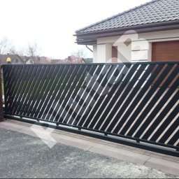 Robstal - Nowoczesna, przesuwna brama wjazdowa z ukośnymi szczeblami, osadzona między ceglanymi słupkami. Widoczny fragment ogrodzenia i domu w tle.