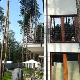 Nowoczesny budynek z balkonami z balustradami. Ujęcie z perspektywy ogrodu z drzewami. Widoczne okna z roletami wewnętrznymi i elementy architektury.