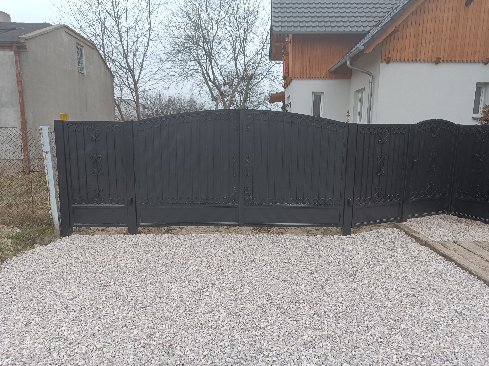 Czarna, metalowa brama wjazdowa z dekoracyjnymi elementami, zamontowana przed domem z jasną elewacją i szarym dachem, na podłożu wysypanym białym kamieniem.