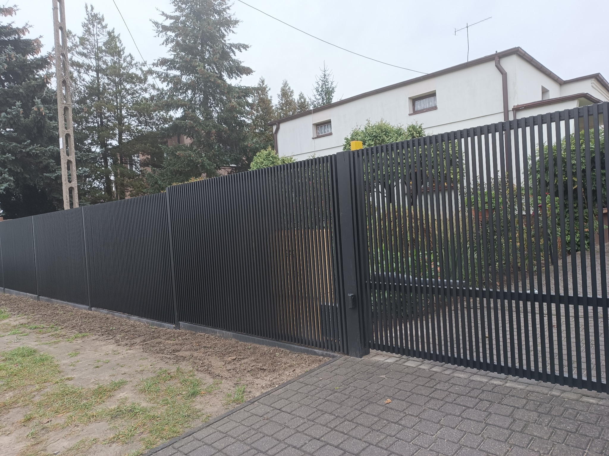 Nowoczesne, grafitowe ogrodzenie panelowe z bramą przesuwną, pionowe lamele. Tło: dom jednorodzinny i drzewa. Minimalistyczny design, estetyczne wykończenie.