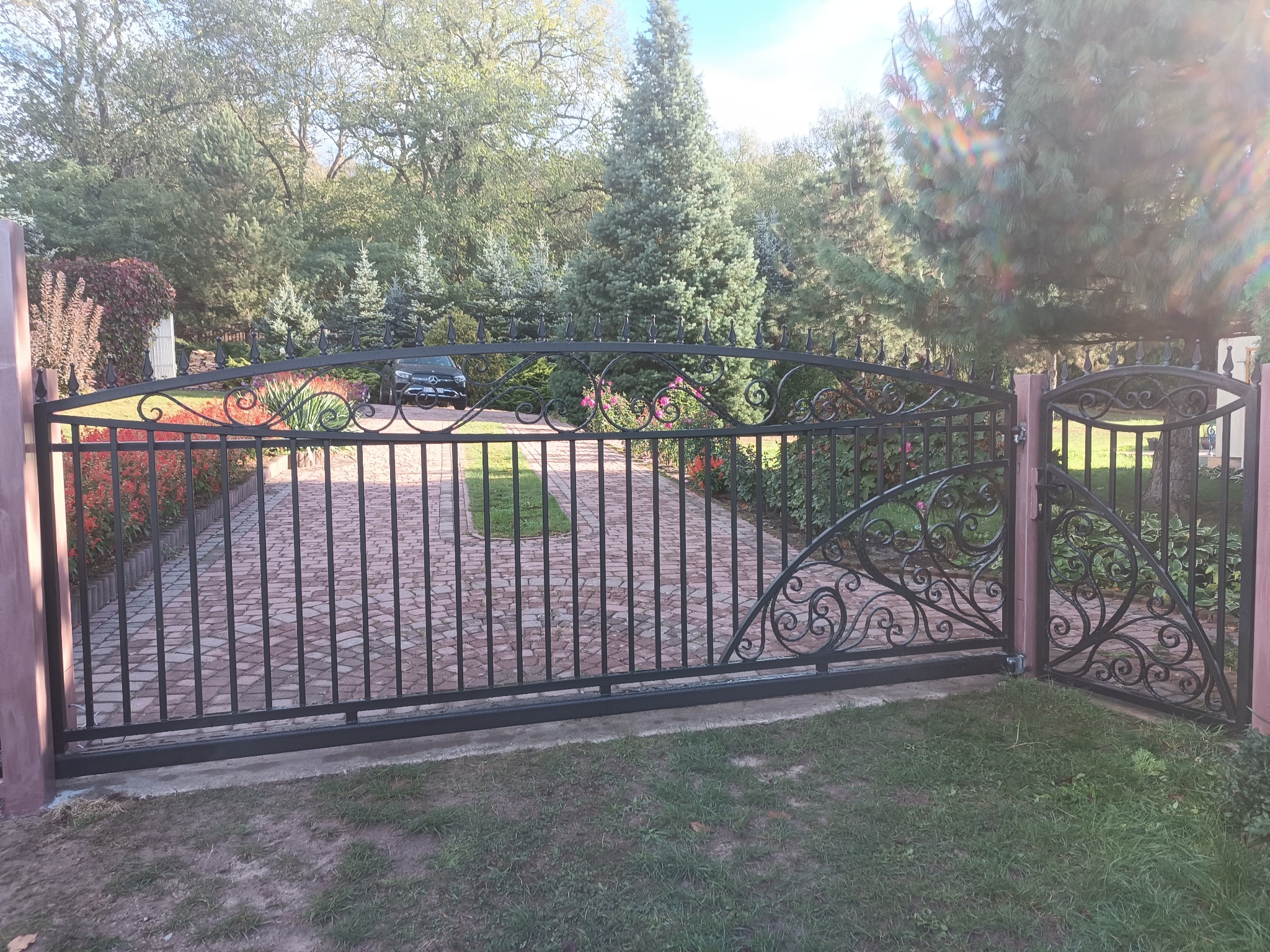 Kuta brama wjazdowa z furtką, zdobiona ornamentami. Na podjeździe kostka brukowa, w tle zieleń drzew i krzewów. W oddali zaparkowany czarny samochód.