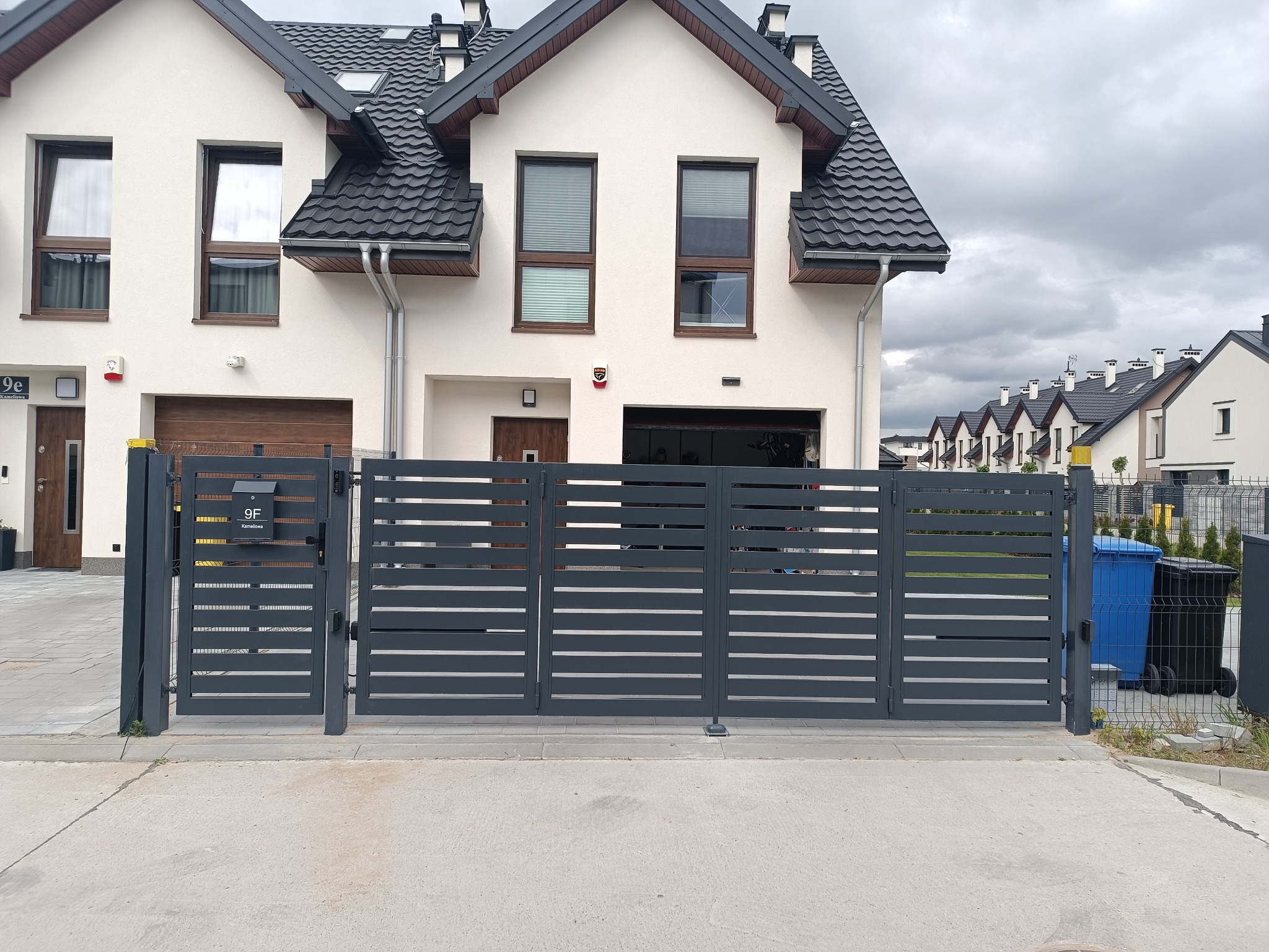 Nowoczesna, pozioma brama wjazdowa w kolorze grafitowym, z numerem domu 9F na skrzynce pocztowej, na tle białego domu z ciemną dachówką i pochmurnego nieba.