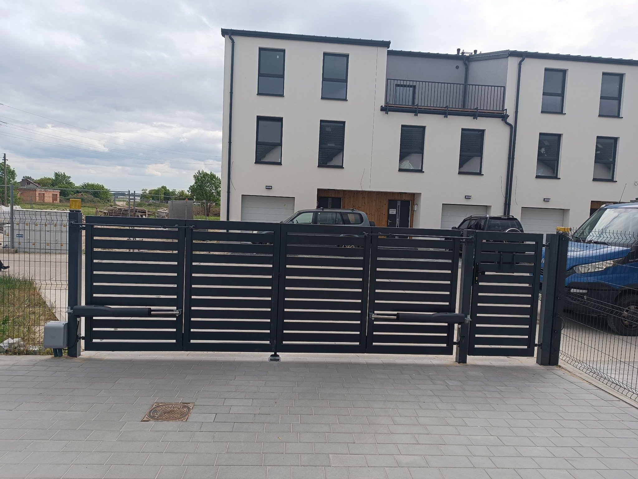 Nowoczesna, przesuwna brama wjazdowa w kolorze grafitowym, z automatyką, na tle szeregu domów jednorodzinnych. Widoczna kostka brukowa i fragment ogrodzenia panelowego.