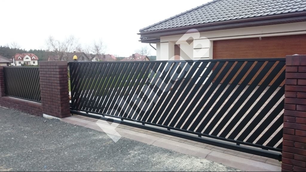 Nowoczesna, przesuwna brama wjazdowa z ukośnymi szczeblami, osadzona między ceglanymi słupkami. Widoczny fragment ogrodzenia i domu w tle.