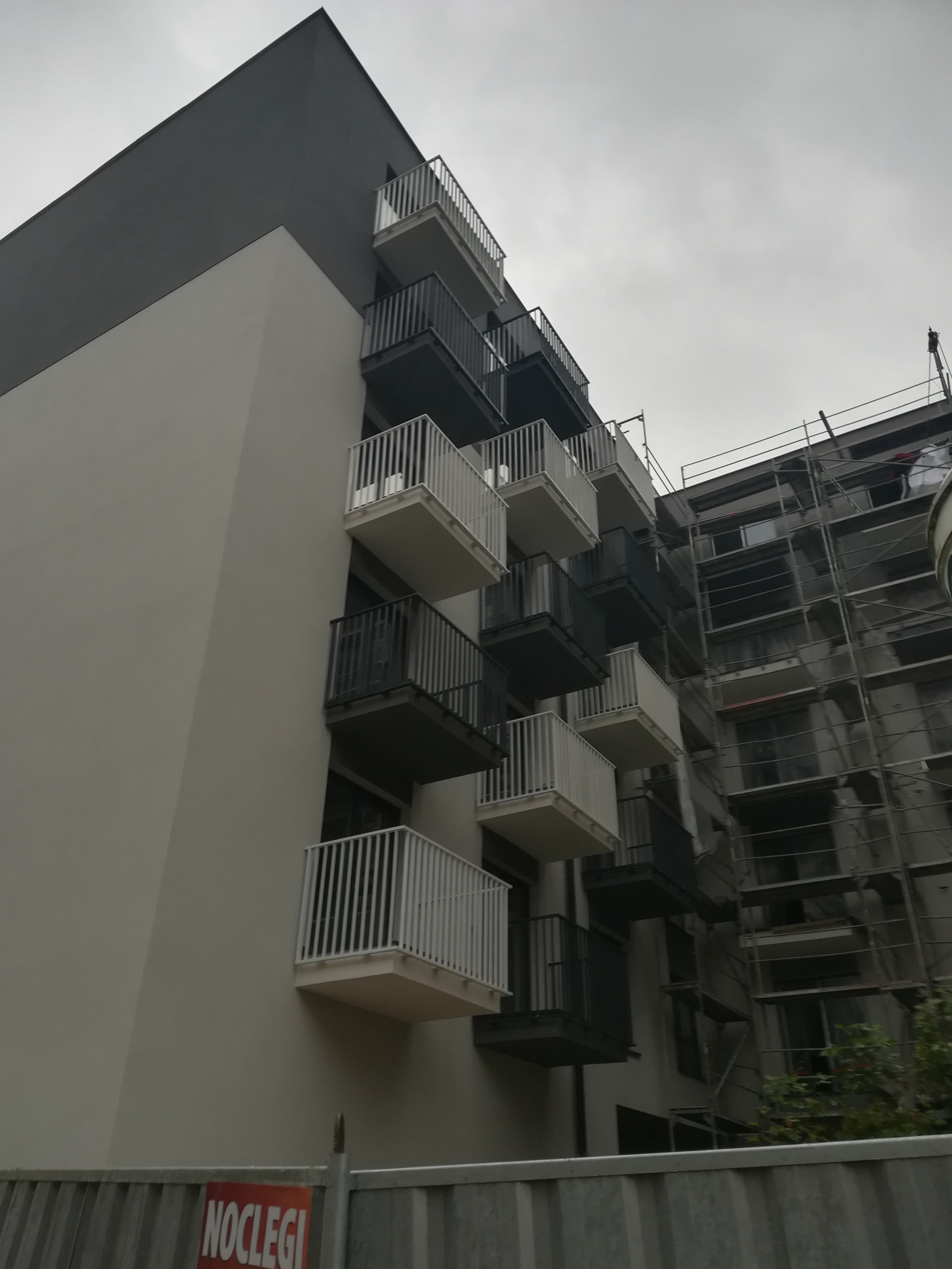 Nowoczesny budynek z balkonami o białych i ciemnych balustradach, widok z dołu na tle pochmurnego nieba. Część budynku w rusztowaniach.
