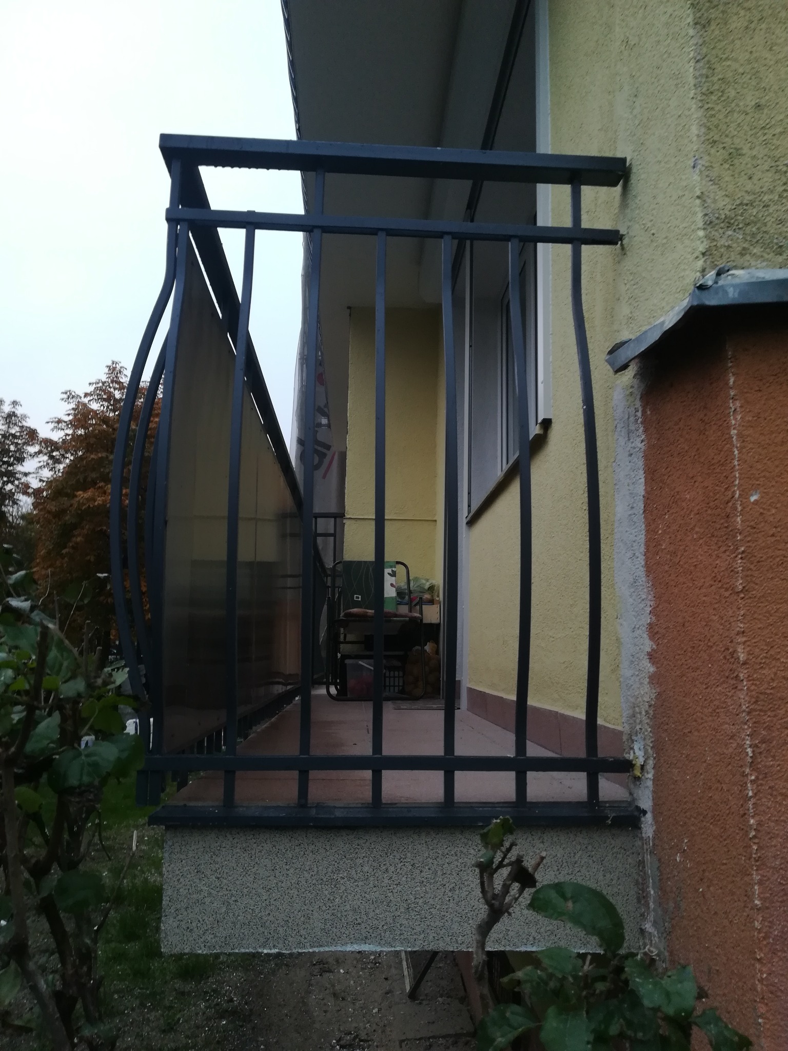 Czarna, metalowa balustrada balkonowa z elementami giętymi, zamontowana na balkonie z widocznym fragmentem elewacji budynku i roślinnością na pierwszym planie.