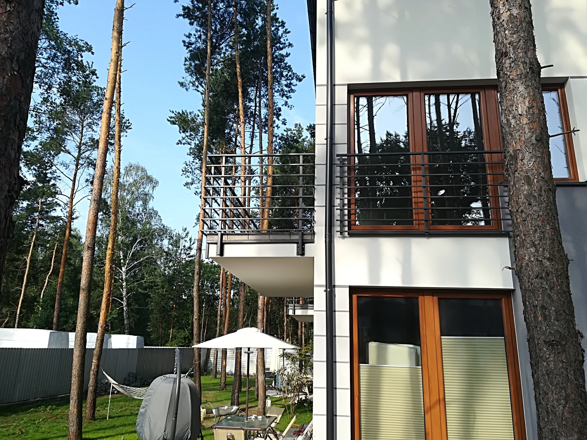 Nowoczesny budynek z balkonami z balustradami. Ujęcie z perspektywy ogrodu z drzewami. Widoczne okna z roletami wewnętrznymi i elementy architektury.