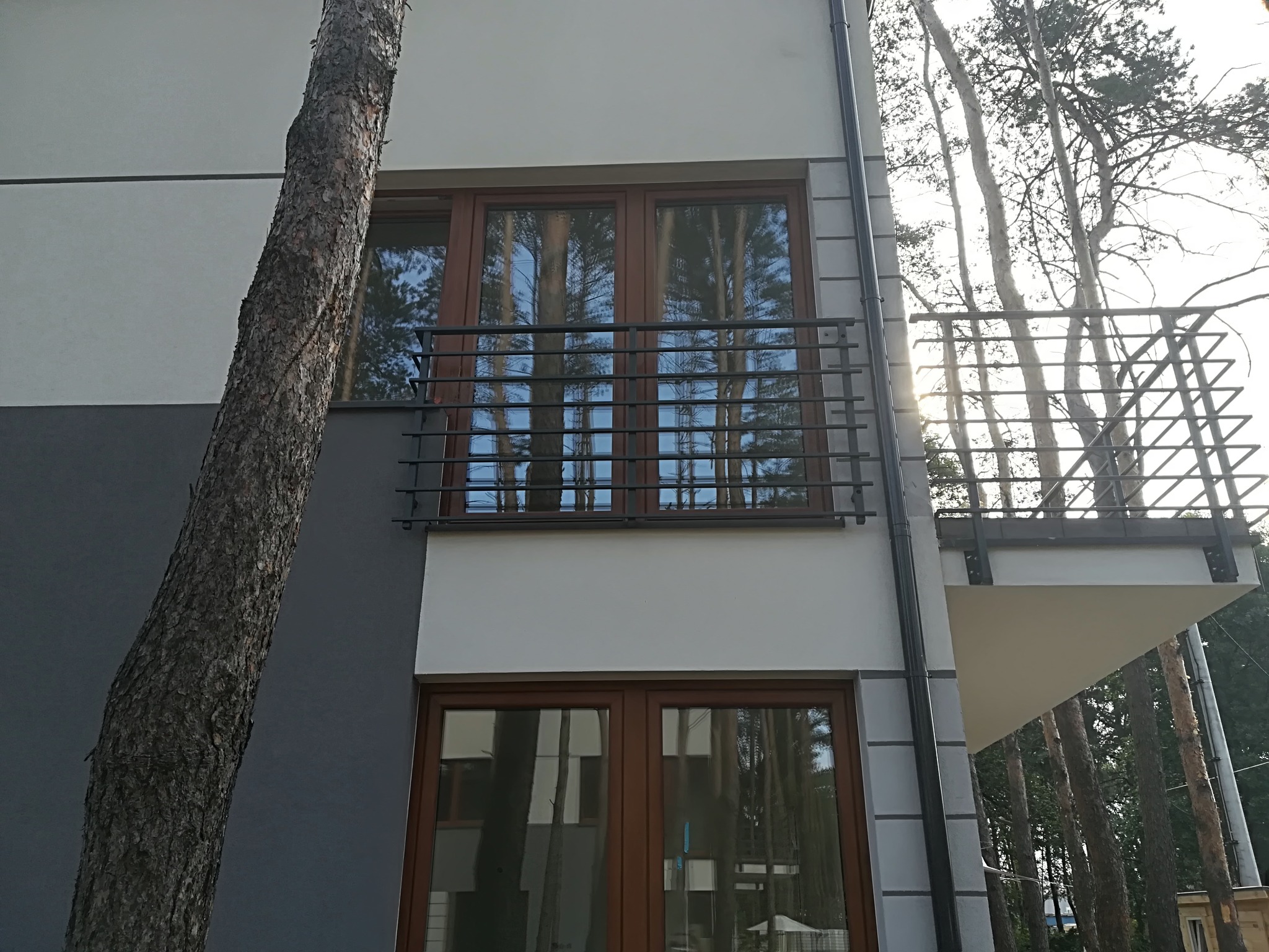 Nowoczesny balkon z metalową balustradą w budynku wielorodzinnym, widok z dołu na tle drzew, z elementami brązowej stolarki okiennej.