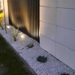 Nowoczesne ogrodzenie z paneli pionowych, oświetlone punktowymi lampami wzdłuż białego żwiru. Elementy betonowe tworzą solidną podstawę, a zieleń dodaje naturalnego charakteru.