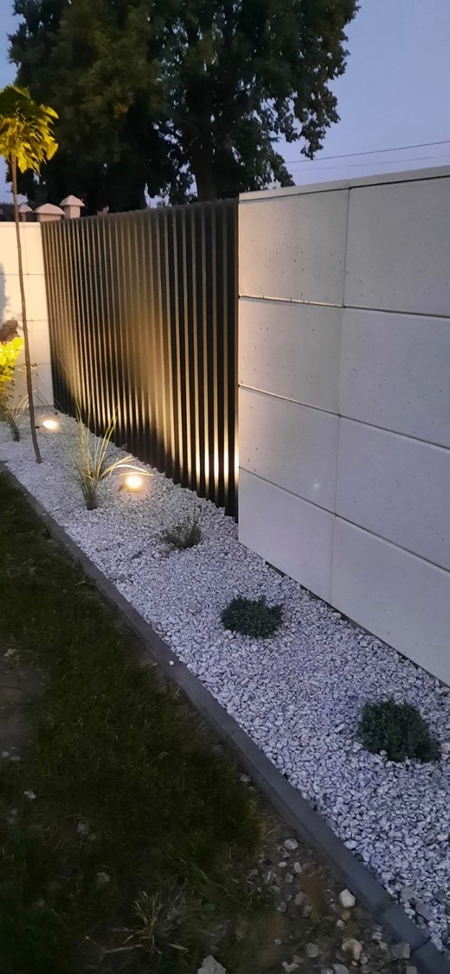 Nowoczesne ogrodzenie z paneli pionowych, oświetlone punktowymi lampami wzdłuż białego żwiru. Elementy betonowe tworzą solidną podstawę, a zieleń dodaje naturalnego charakteru.