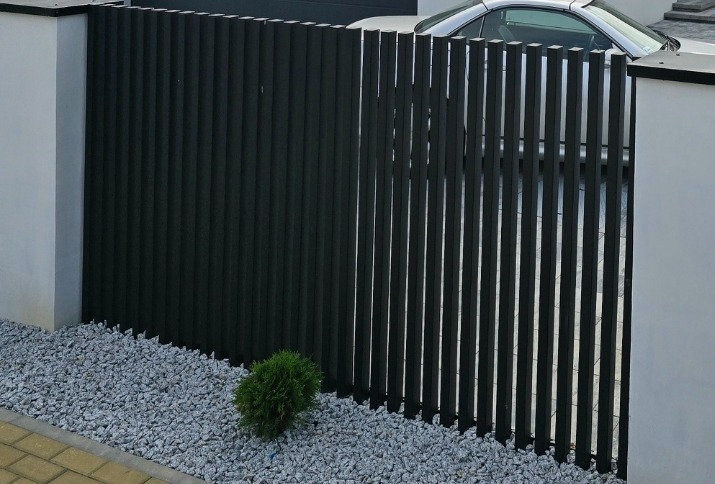 Ciemne, pionowe panele ogrodzeniowe z widocznym samochodem w tle, otoczone białymi słupkami i szarym żwirem. Niewielka, okrągła roślina u podstawy ogrodzenia.