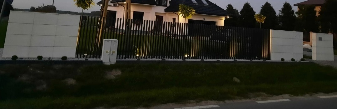 Nowoczesne ogrodzenie z czarnych, pionowych listew i białych, kwadratowych paneli betonowych, oświetlone punktowymi lampami wzdłuż podstawy. Dom w tle.