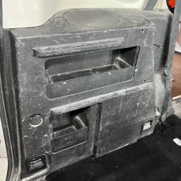 Lucky Detailing Polska OLEKSANDR SHCHUTSKYI - Brudny, czarny panel boczny w bagażniku samochodu z widocznymi schowkami i głośnikiem, wymagający czyszczenia. Widoczne ślady użytkowania i zabrudzenia.