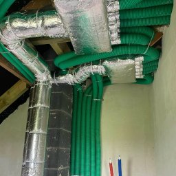 ZHU Wojciech Szczęsny - Zielone rury wentylacyjne i izolowane srebrną folią kanały w budynku, zamocowane do sufitu i ściany. Elementy instalacji rekuperacji.