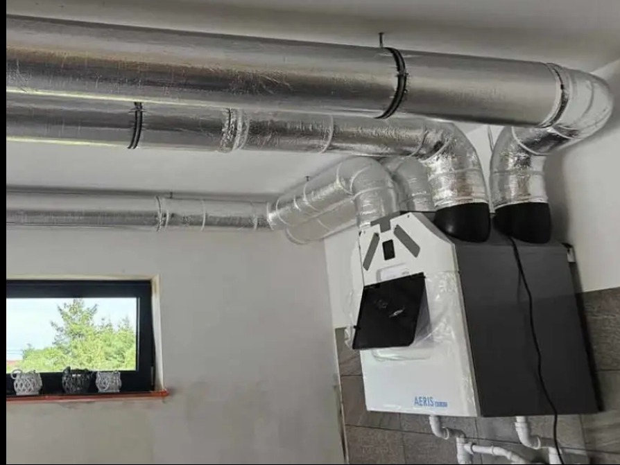 Nowoczesna instalacja rekuperacji AERIS zamontowana przy ścianie, z widocznymi srebrnymi rurami wentylacyjnymi pod sufitem. Okno z dekoracjami w tle.