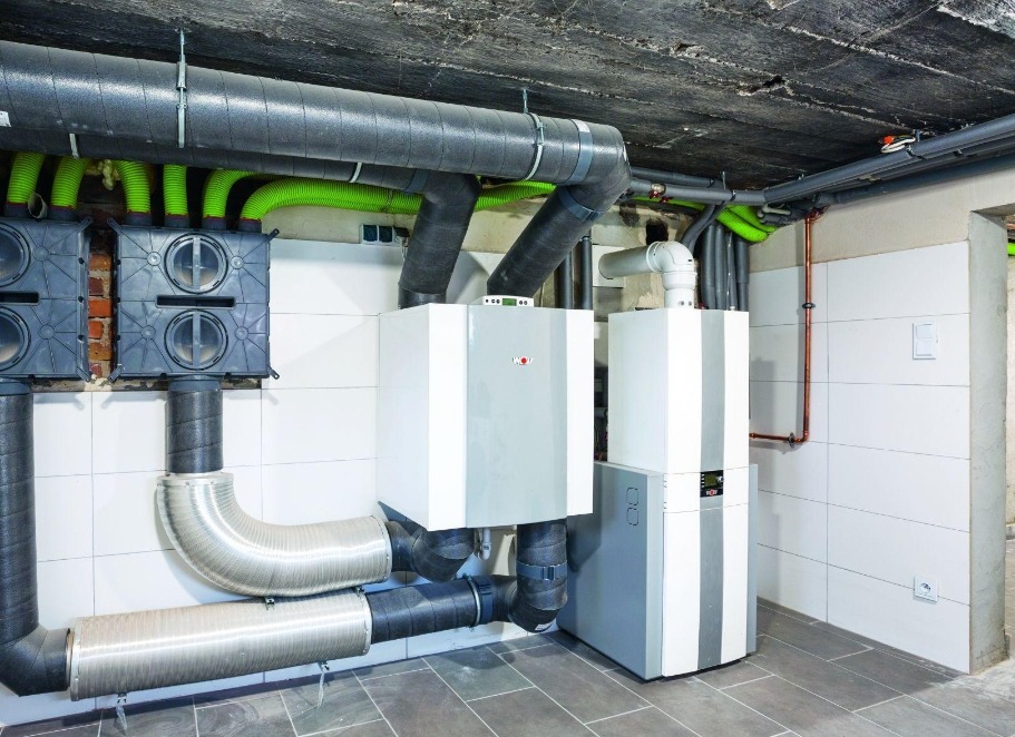Zainstalowana rekuperacja w pomieszczeniu technicznym: szare rury wentylacyjne, centrale rekuperacyjne Wolf, białe płytki na ścianach i szara podłoga.