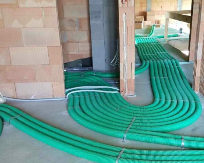 Zielone rury wentylacyjne ułożone równolegle na szarej posadzce w surowym wnętrzu budynku z ceglanymi ścianami. Widoczne mocowania rur.